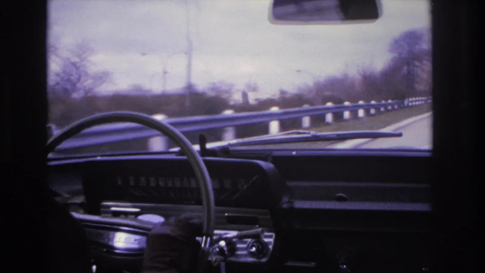 Driver's perspective in what appears to be a 1960s American car, showing the dark instrument panel and steering wheel with a guardrail-lined highway and utility poles visible ahead, circa 1966.