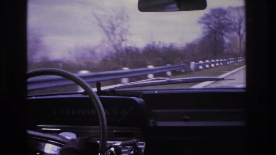 Forward windshield view from what appears to be a 1966 car on a northeastern US highway, showing a prominent metal guardrail, bare winter trees on both sides, and an overcast sky.