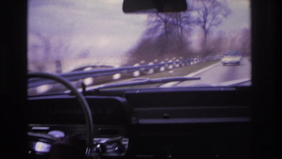 Windshield POV from a 1960s car on a highway, possibly in New Jersey circa 1966, showing a white vehicle ahead, metal guardrail, bare winter trees, and an overcast sky.