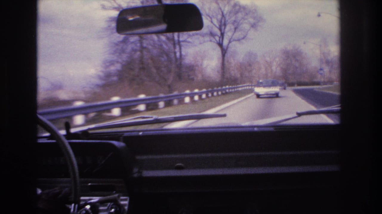 8mm home movie frame circa 1966 showing driver's-eye view of a divided New Jersey highway with metal guardrail, bare trees, and 1960s-era cars ahead.