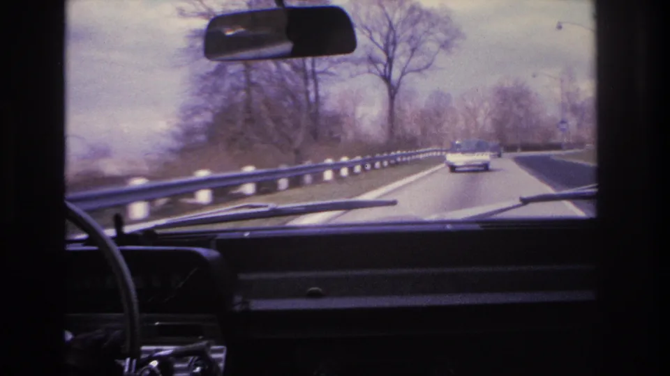 8mm home movie frame circa 1966 showing driver's-eye view of a divided New Jersey highway with metal guardrail, bare trees, and 1960s-era cars ahead.