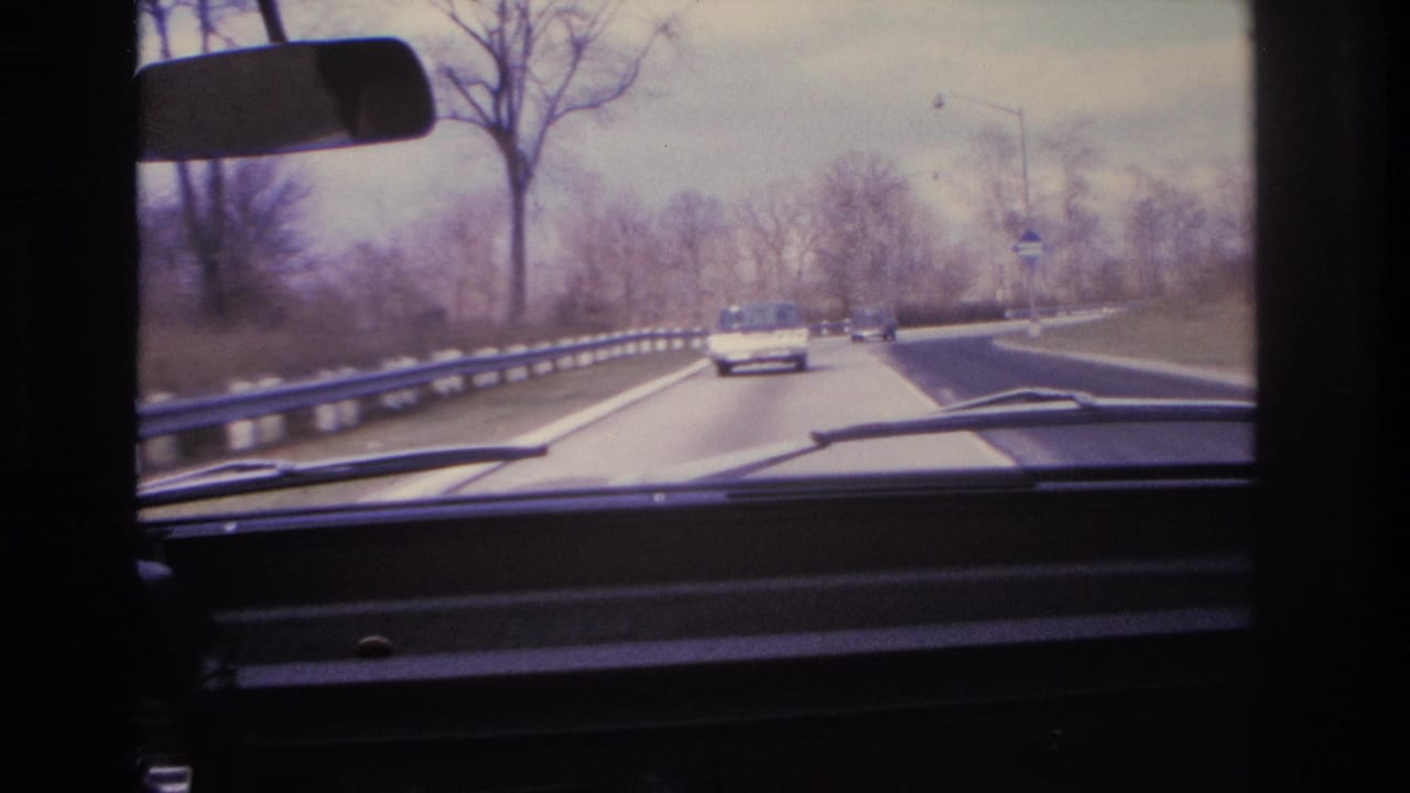8mm home movie frame circa 1966 showing a continuing windshield drive along a divided highway with guardrail, bare trees, and a period car ahead.
