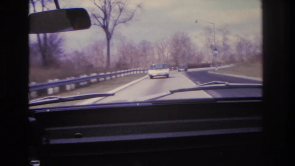 8mm home movie frame circa 1966 showing a continuing windshield drive along a divided highway with guardrail, bare trees, and a period car ahead.