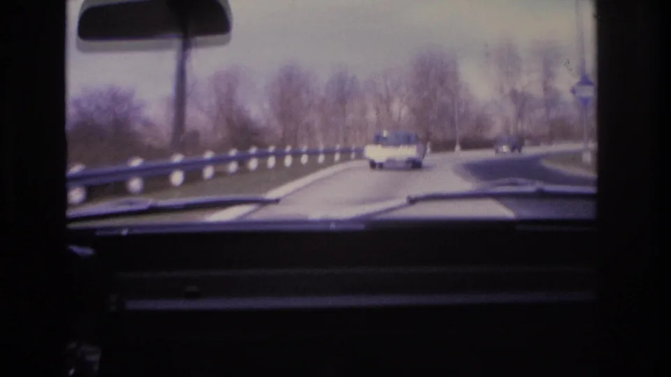 8mm home movie frame circa 1966 showing a highway curve from the driver's perspective, with guardrail, bare trees, and a light-colored 1960s vehicle ahead.