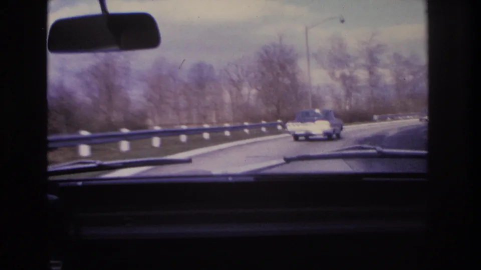 8mm home movie frame circa 1966 showing a divided highway with guardrail, utility pole, bare trees, and a 1960s-era car ahead from the driver's perspective.