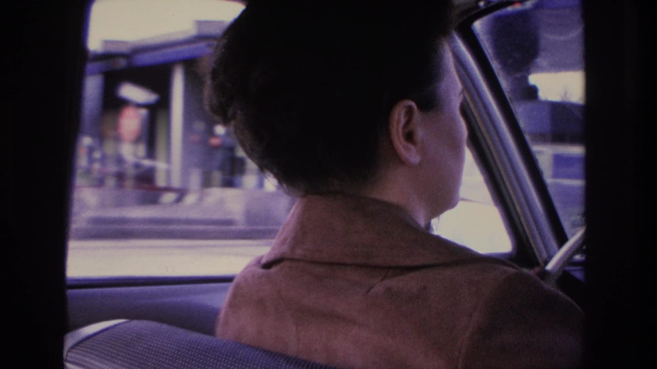 Woman in brown jacket at the steering wheel of a 1960s-era car, filmed from the backseat, with urban Hoboken streetscape blurred through the side window, 1966.