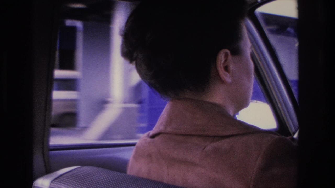 Woman at steering wheel seen from behind, large pale-walled building passing through side window.
