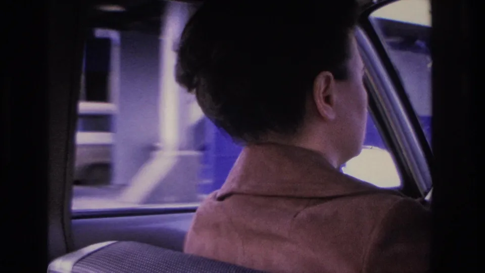 Woman driving past a large pale building on a Hoboken street, filmed on 8mm from the backseat, consistent with 1966 urban NJ streetscape.