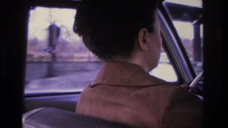 Backseat 8mm shot of woman driving past bare trees and a utility pole on a Hoboken area street, 1966.