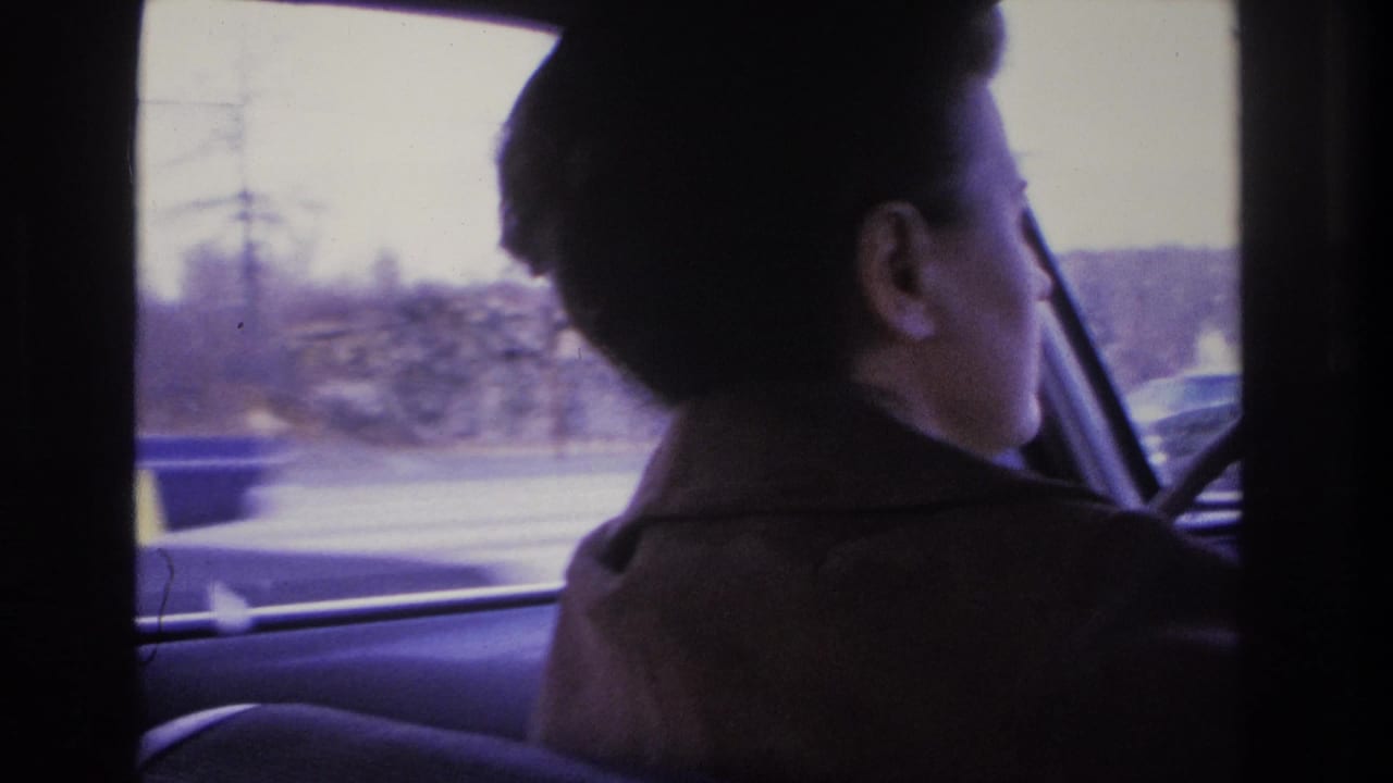 A woman with short dark hair and a dark coat drives, photographed from the rear seat in 8mm footage circa 1966, suburban landscape and bare trees visible through the side window.