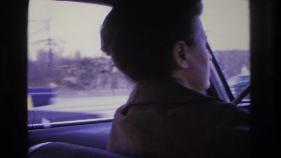 A woman with short dark hair and a dark coat drives, photographed from the rear seat in 8mm footage circa 1966, suburban landscape and bare trees visible through the side window.