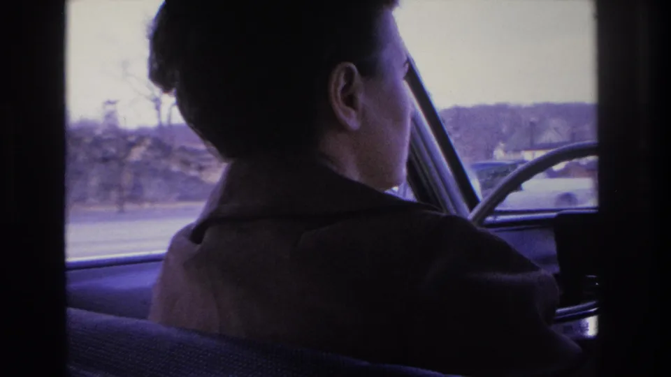 A woman with short styled dark hair and a dark coat, shot from the right rear seat in 8mm home movie footage circa 1966, driving through a suburban area with bare winter trees beyond the window.