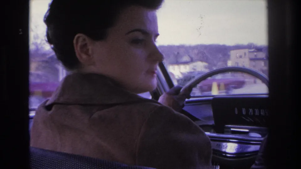 Partial profile of a woman at the wheel, steering wheel and chrome dashboard elements in view, shot in 8mm circa 1966, with colorful roadside structures glimpsed through the side window.