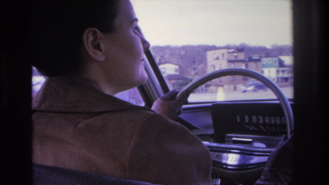 Young woman in a brown jacket driving a mid-1960s car, seen in profile from the passenger seat; residential buildings through windshield, 1966 8mm home movie, Hoboken, NJ.