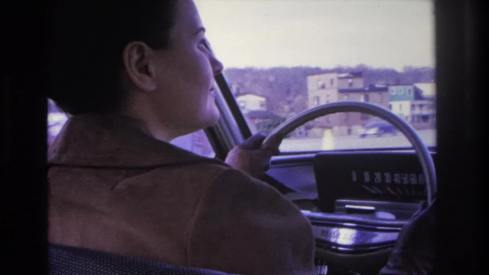 Young woman in a brown jacket driving a mid-1960s car, seen in profile from the passenger seat; residential buildings through windshield, 1966 8mm home movie, Hoboken, NJ.