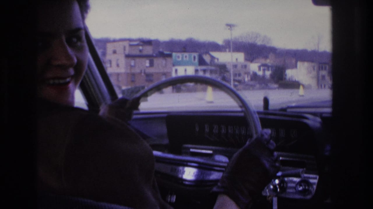 Young woman with hair in a bun turns to smile at the camera from the driver's seat; urban streetscape through windshield, 1966 8mm home movie, Hoboken, NJ.