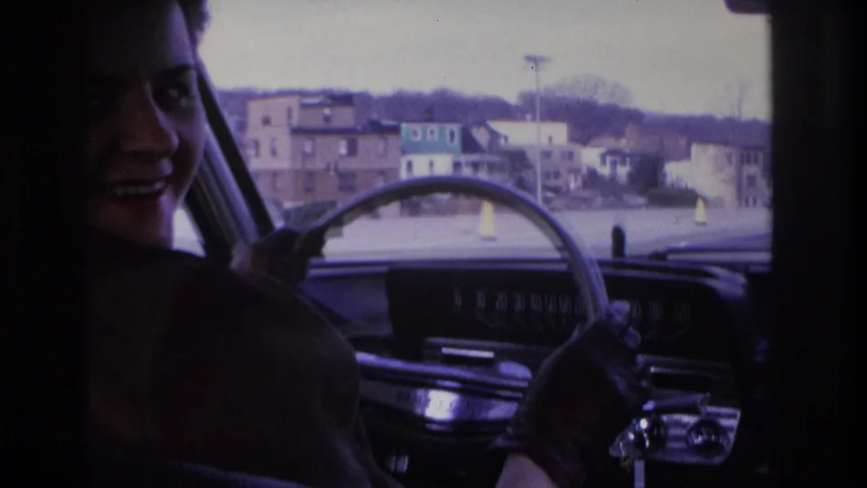 Young woman with hair in a bun turns to smile at the camera from the driver's seat; urban streetscape through windshield, 1966 8mm home movie, Hoboken, NJ.