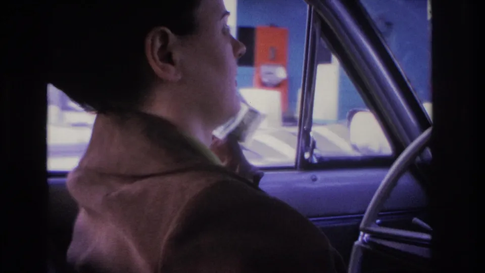 Young woman with dark hair in a bun driving, viewed from behind; colorful commercial buildings or signage through the side window, 1966 8mm home movie, Hoboken, NJ area.