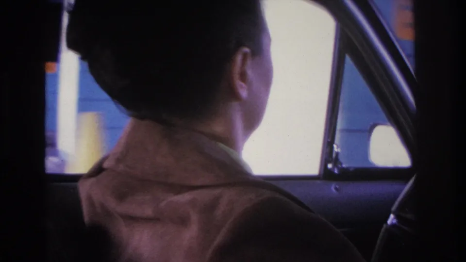 Young woman in brown jacket viewed from behind the driver's seat; blue and yellow commercial structure through the window, 1966 8mm home movie, Hoboken, NJ area.