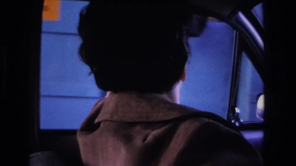 1966 8mm home movie, New Jersey: woman driver in brown wool coat viewed from rear, cool blue ambient light through car window.
