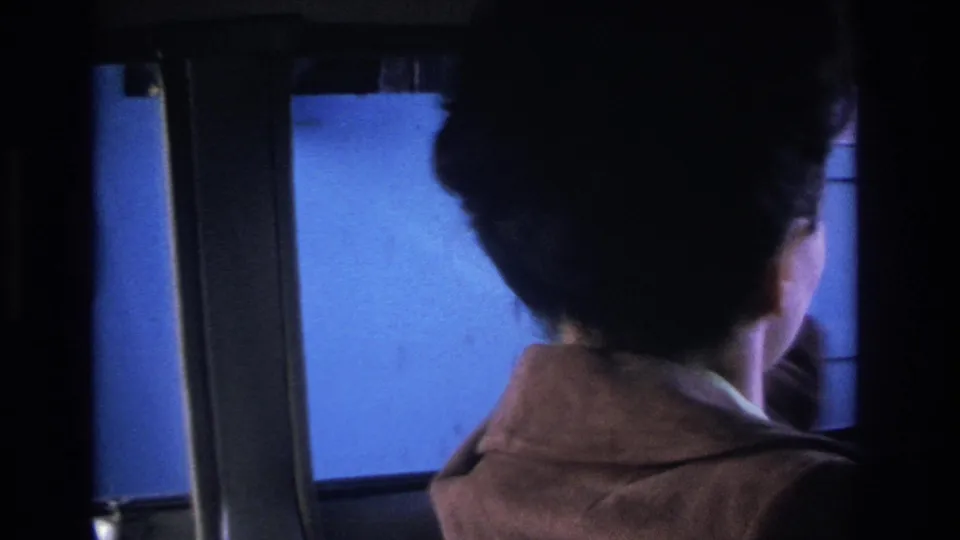 1966 8mm footage: woman driver in brown coat seen from rear inside vintage car, cool blue ambient light after toll payment.