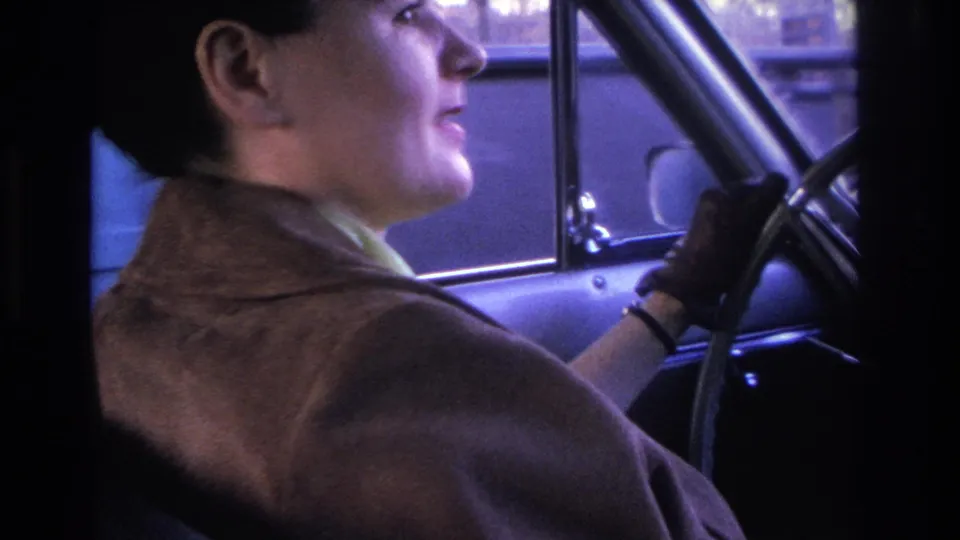 1966 8mm home movie: woman driver in profile in brown wool coat and dark gloves, slight smile, vintage steering wheel and dashboard, blue-lit window.