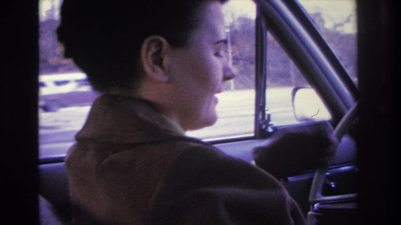 Dark-haired woman in brown coat in right-facing profile, blurry suburban street and structures visible through car window; consistent with 1966 Hoboken, New Jersey home movie.