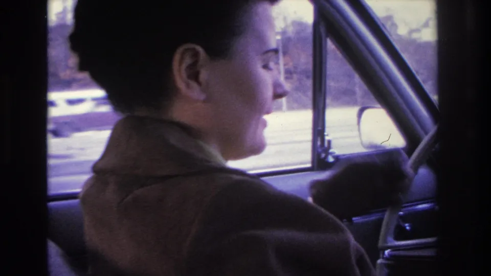 Dark-haired woman in brown coat in right-facing profile, blurry suburban street and structures visible through car window; consistent with 1966 Hoboken, New Jersey home movie.