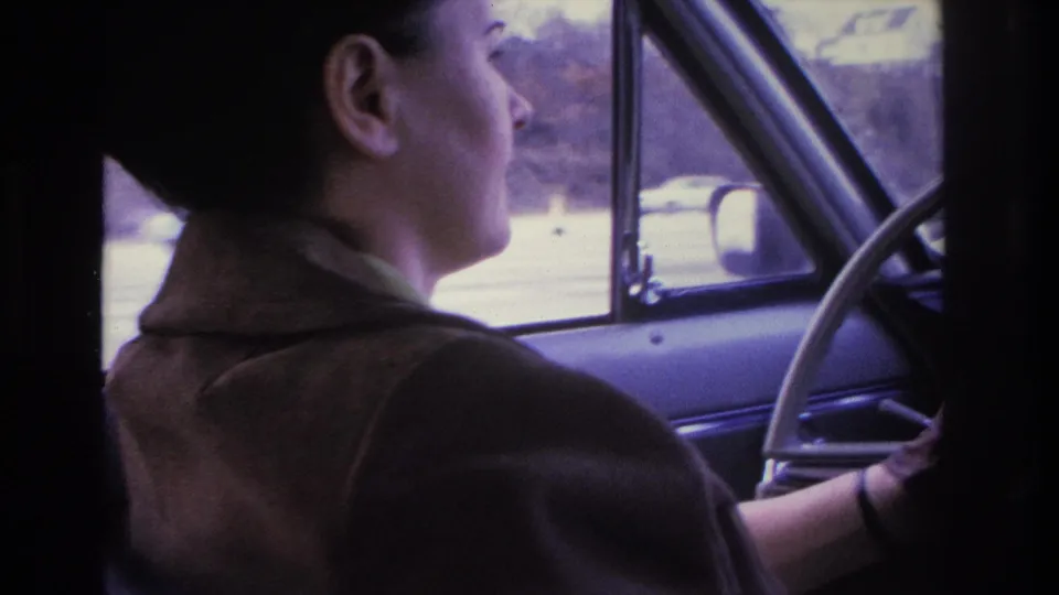Dark-haired woman in brown coat gripping a thin-rimmed steering wheel, shot from passenger seat; blurry street scene visible through window, consistent with a 1960s American car interior.