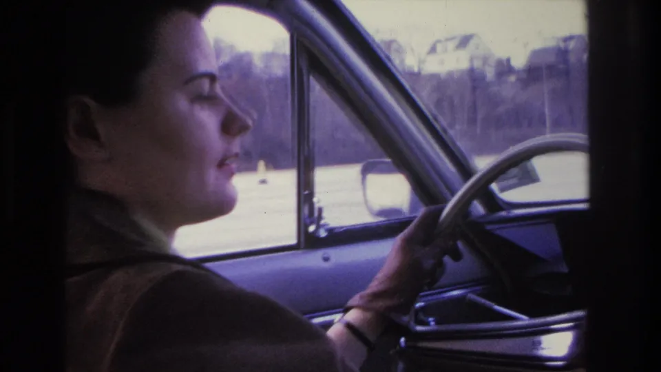 Dark-haired woman in brown coat at the steering wheel, appearing to smile or talk, wristwatch on wrist visible; blurry suburban street through window consistent with 1966 New Jersey home movie.