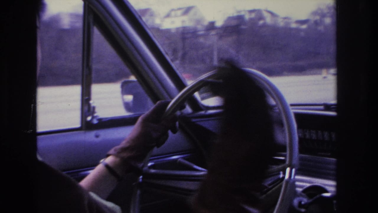 1966 Hoboken home-movie frame showing a woman at the wheel of a car, her hand near a cigarette, with road and houses through the windshield.