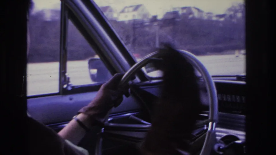 1966 Hoboken home-movie frame showing a woman at the wheel of a car, her hand near a cigarette, with road and houses through the windshield.