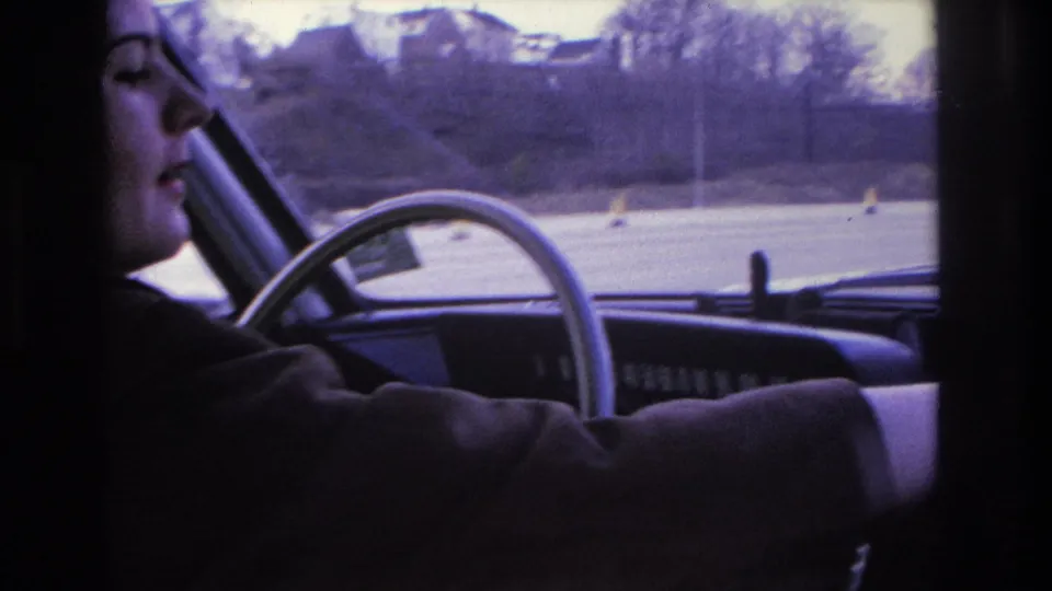 1966 Hoboken home-movie frame showing the woman driver in profile, with the steering wheel, dashboard, road, and houses visible through the windshield.