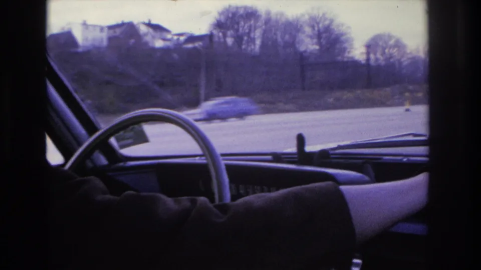 1966 Hoboken frame from inside the car, showing the driver’s arm reaching across the dashboard as the road and a passing car appear outside.