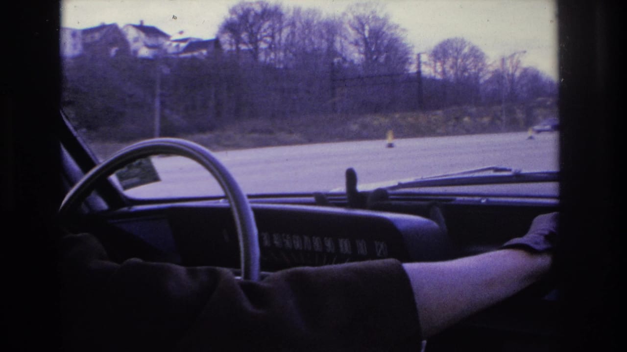 1966 Hoboken-area home-movie frame from inside a car, showing a steering wheel, dashboard, driver arm, roadway cones, and bare trees beyond the windshield.