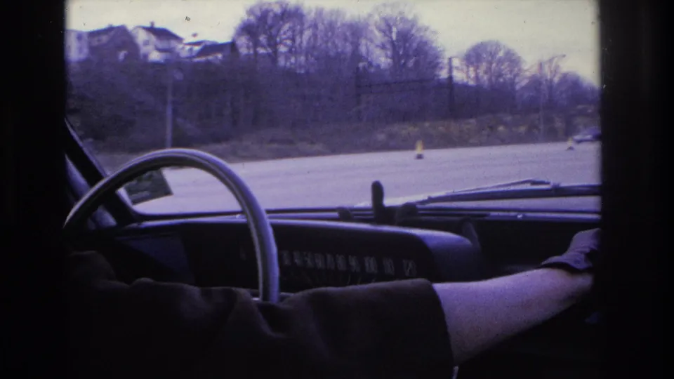 1966 Hoboken-area home-movie frame from inside a car, showing a steering wheel, dashboard, driver arm, roadway cones, and bare trees beyond the windshield.