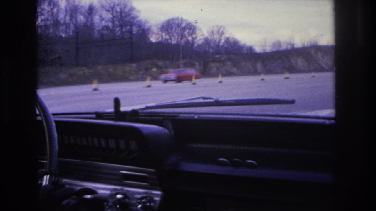 Windshield view from a car with dashboard, wipers, cones, a passing red vehicle, and bare roadside trees.