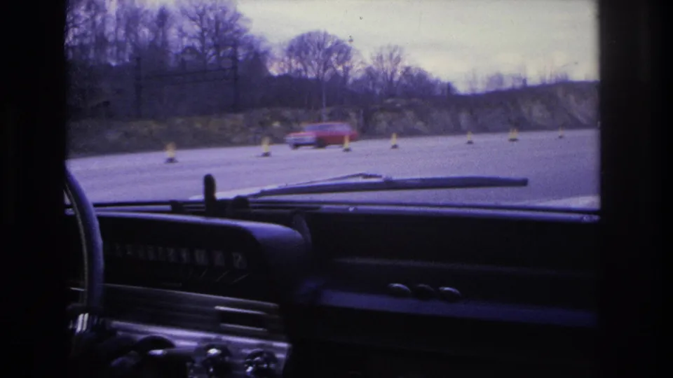 1966 Hoboken-area driving POV with dashboard and windshield wipers in the foreground and a red car passing near traffic cones on a wide road.