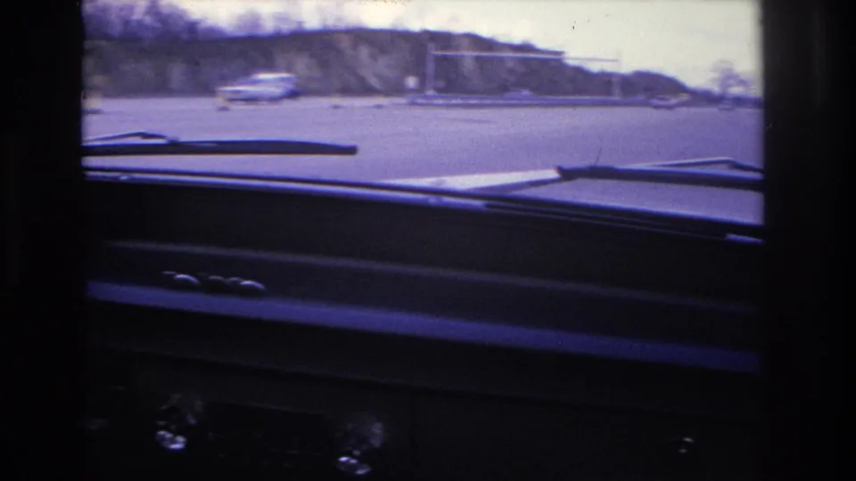 Interior 8mm car view in Hoboken, 1966, looking past the dashboard and wipers toward traffic cones, a passing vehicle, and a low roadside building.