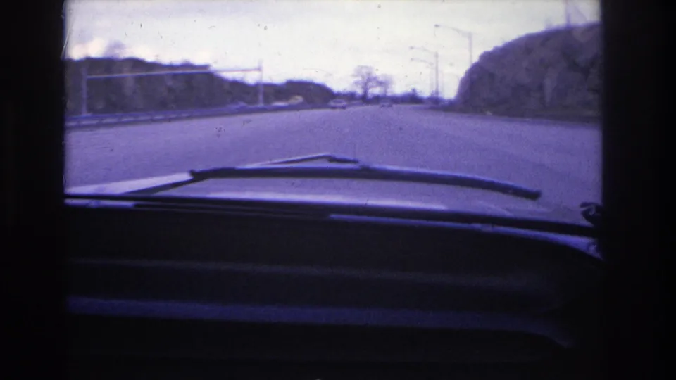 1966 Hoboken-area road view from inside a car, with windshield wipers and dashboard in the foreground and an open roadway, poles, trees, and hillside ahead.