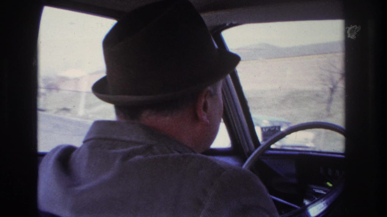 1966 Hoboken home-movie frame of a fedora-wearing driver at the wheel with pale roadside landscape through the windshield.