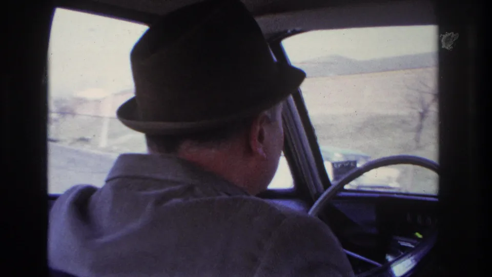 1966 Hoboken home-movie frame of a fedora-wearing driver at the wheel with pale roadside landscape through the windshield.