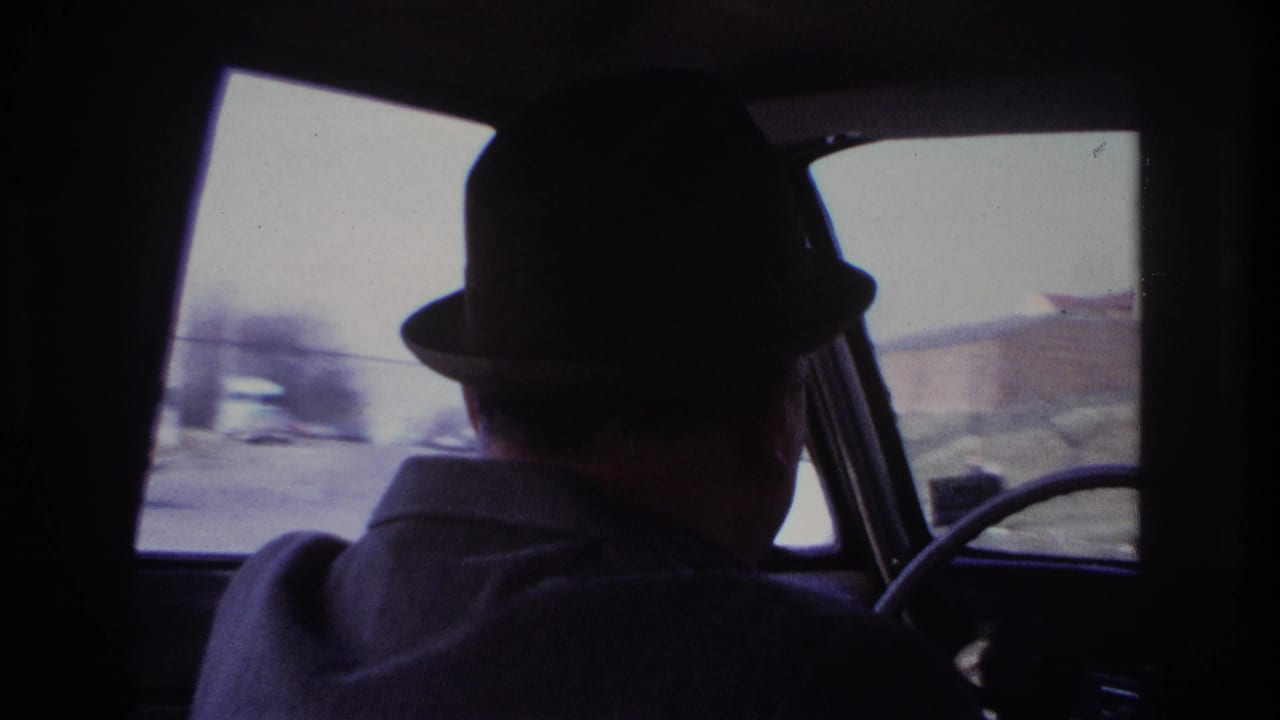 1966 Hoboken home-movie frame showing an older man in a dark fedora driving, with blurred street traffic visible through the car windows.
