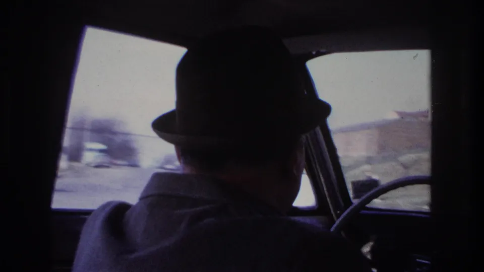 1966 Hoboken home-movie frame showing an older man in a dark fedora driving, with blurred street traffic visible through the car windows.