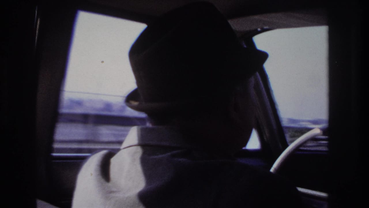 Close back-seat view of the fedora-wearing driver in a 1966 Hoboken home movie, with the roadway and buildings blurred through the windows.