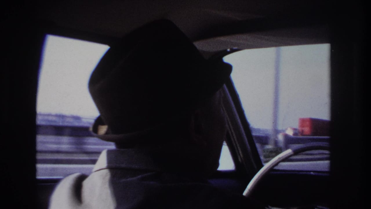 1966 Hoboken home-movie frame showing a silhouetted male driver in a fedora, viewed from the back seat of a moving car.