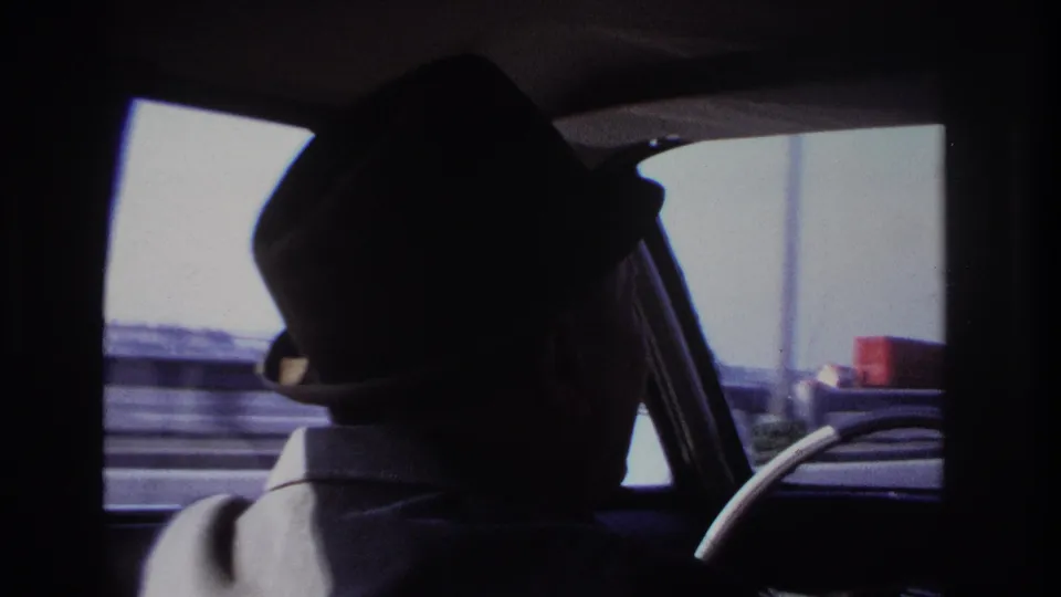 1966 Hoboken home-movie frame showing a silhouetted male driver in a fedora, viewed from the back seat of a moving car.