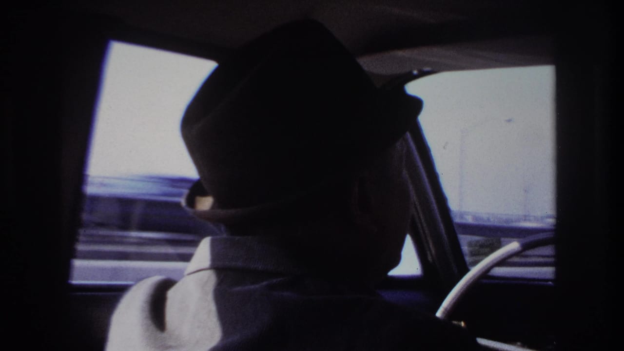 Backseat passenger view of a fedora-wearing driver in a moving car, with Hoboken roadside barriers blurred outside.