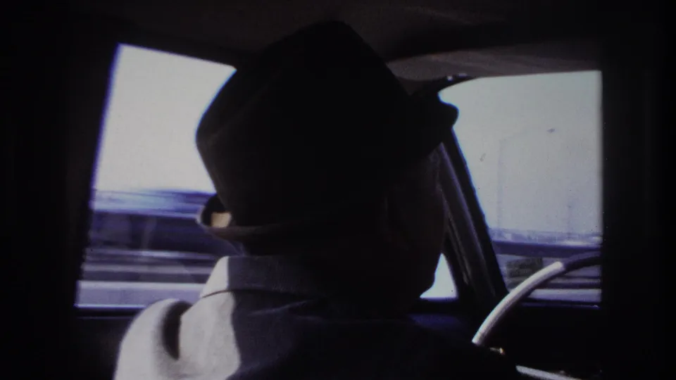 Backseat passenger view of a fedora-wearing driver in a moving car, with Hoboken roadside barriers blurred outside.