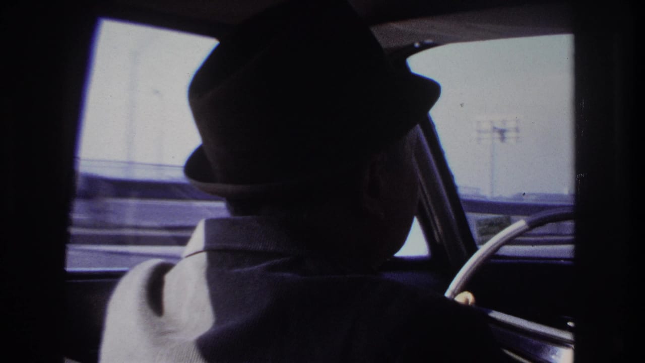 1966 home-movie view from inside a car, showing the driver’s hat and shoulder against a bright Hoboken roadside window.